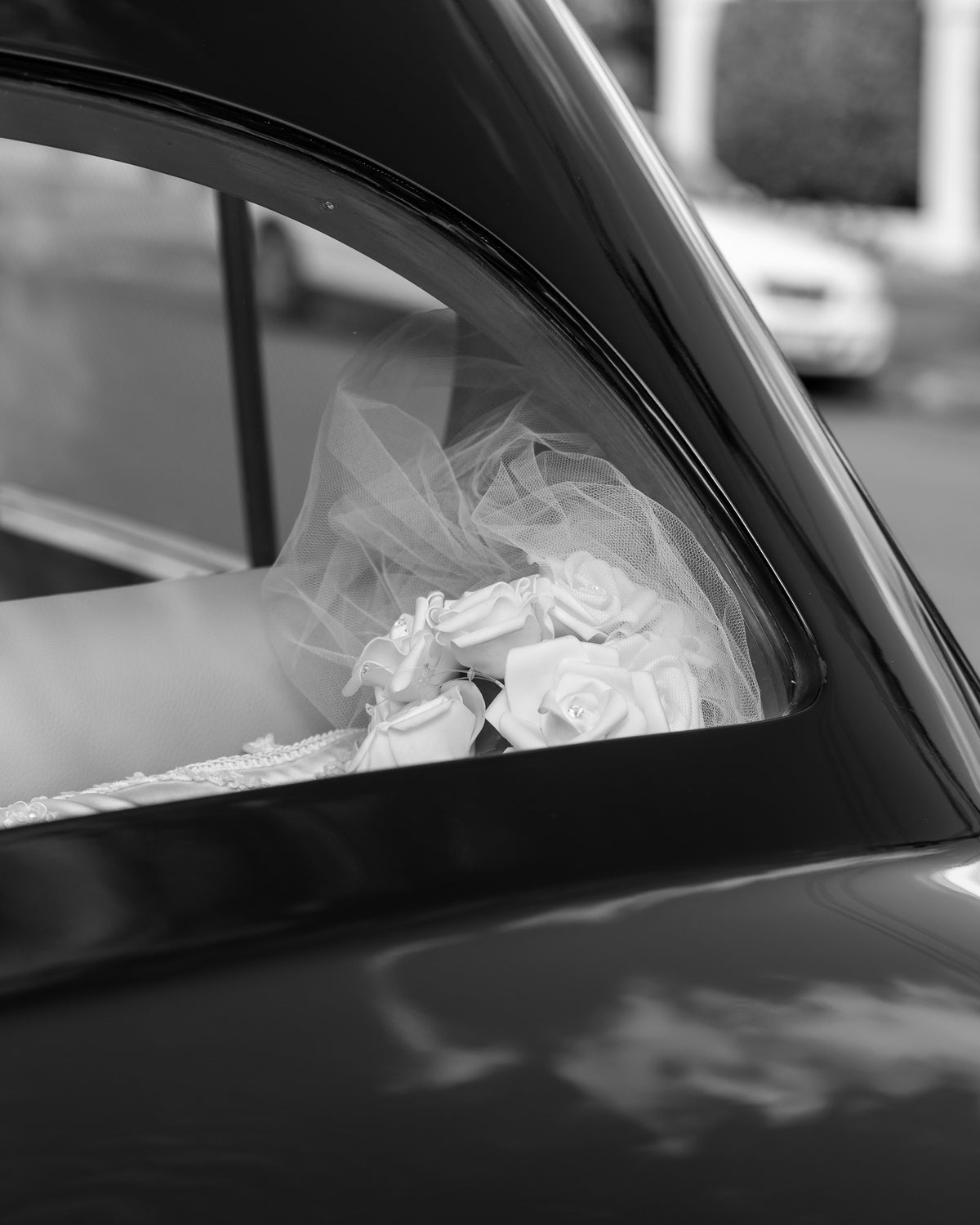Black and white detail of a bridal bouquet and veil beside a car window.