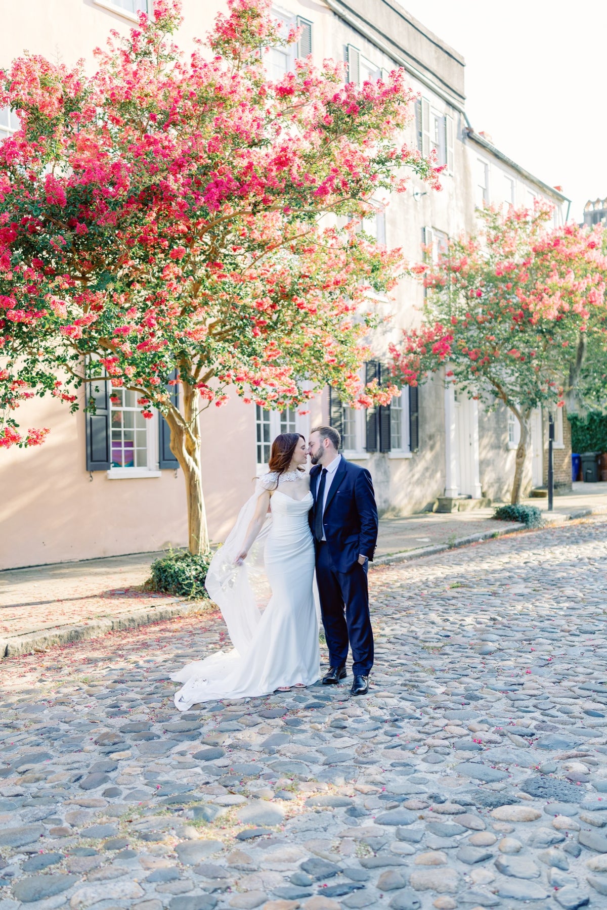 A couple on a Charleston cobblestone street beneath flowering trees. Jolie Connor Photography.