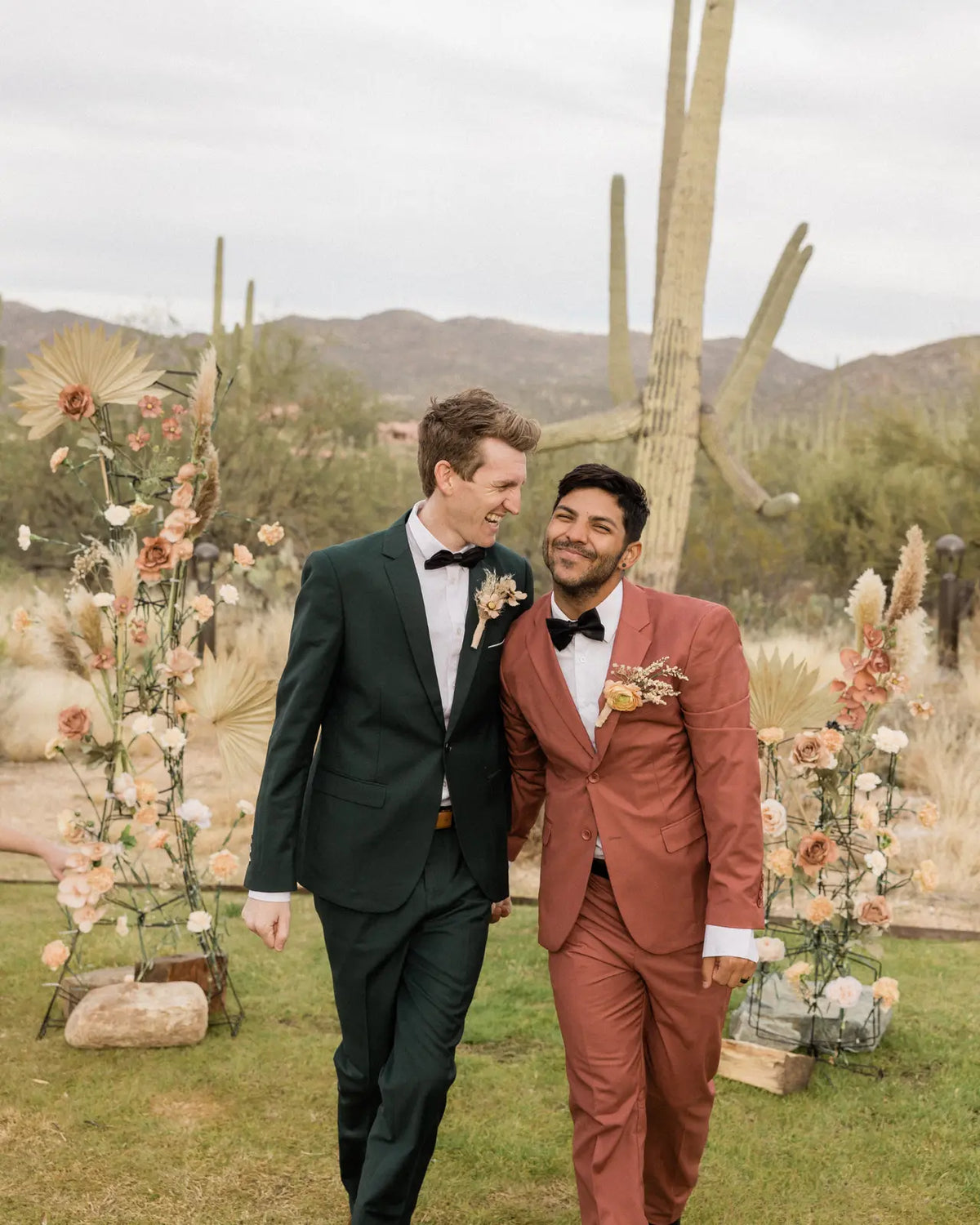Two newlyweds in green and terracotta suits embrace beside their ceremony flowers.