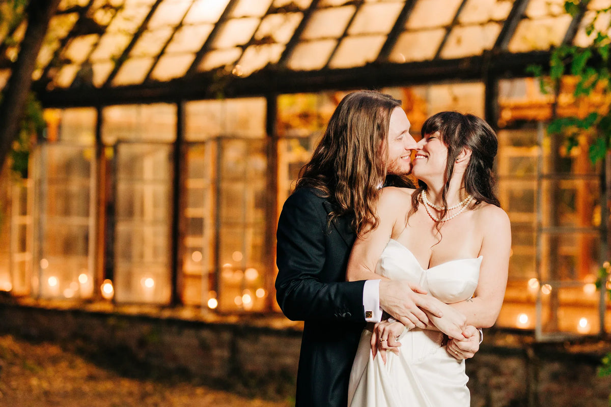 A couple embraces in front of a warmly lit greenhouse.